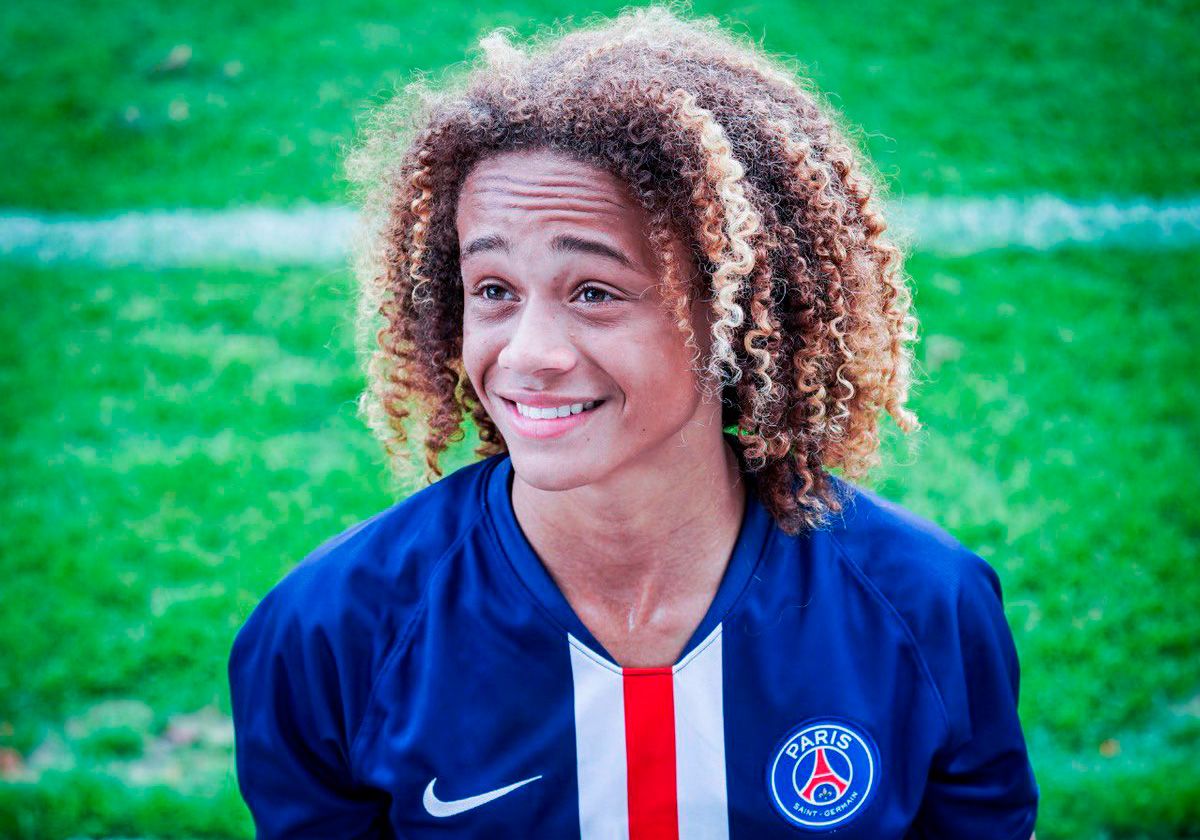  A young male football player with long curly hair is smiling while looking away from the camera and wearing a blue and red jersey with the logo of Paris Saint-Germain.